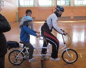 タンデム自転車の前席に大人、後席にアイマスクをした子供が乗っています