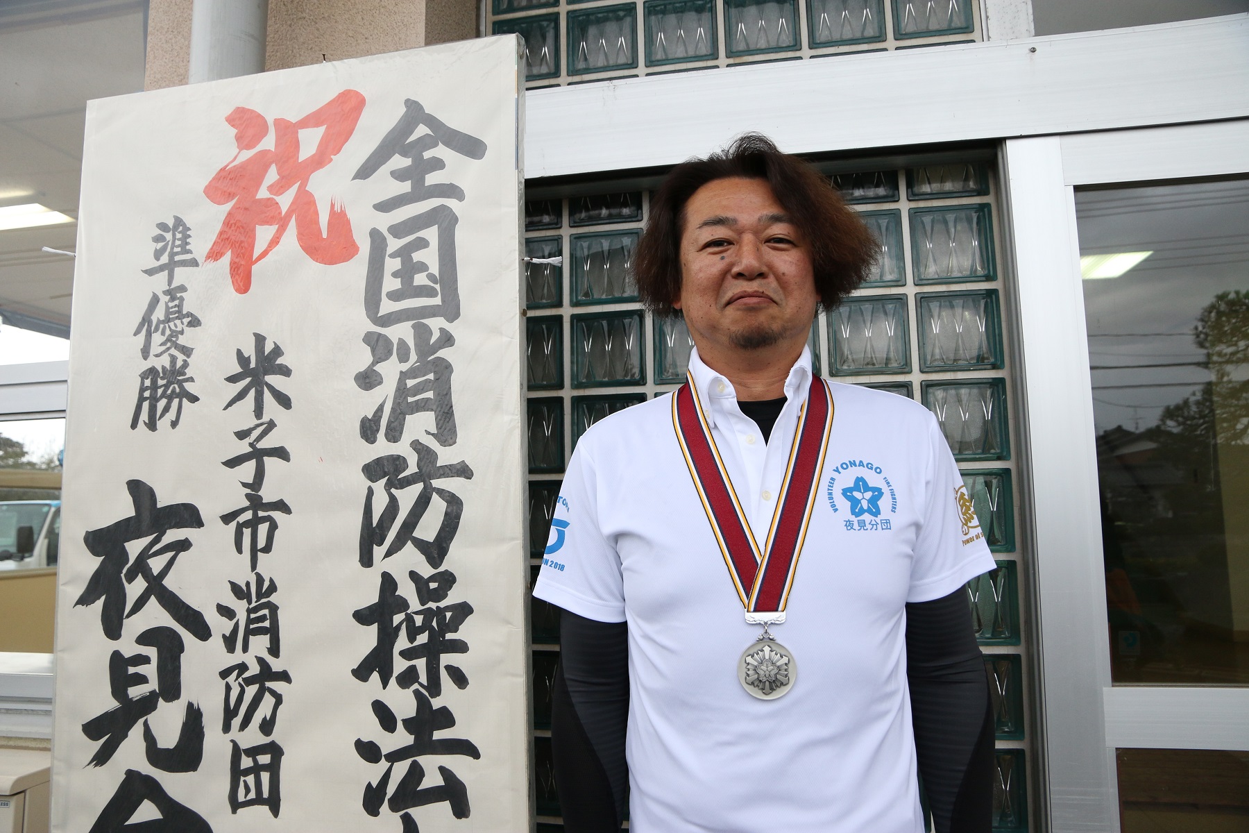 地域の方々の手作り看板の前でメダルをかける西田さん