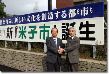 写真：米子市役所前に設置された合併決定を知らせる看板の前で握手する野坂米子市長と田口淀江町長